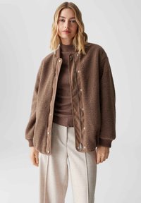 Bruine fleece jas met drukknoopsluiting en een contrasterende stoffen bies. Heeft een losse schouderlijn, ribgebreide manchetten en een ontspannen pasvorm.