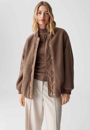 Bruine fleece jas met drukknoopsluiting en een contrasterende stoffen bies. Heeft een losse schouderlijn, ribgebreide manchetten en een ontspannen pasvorm.