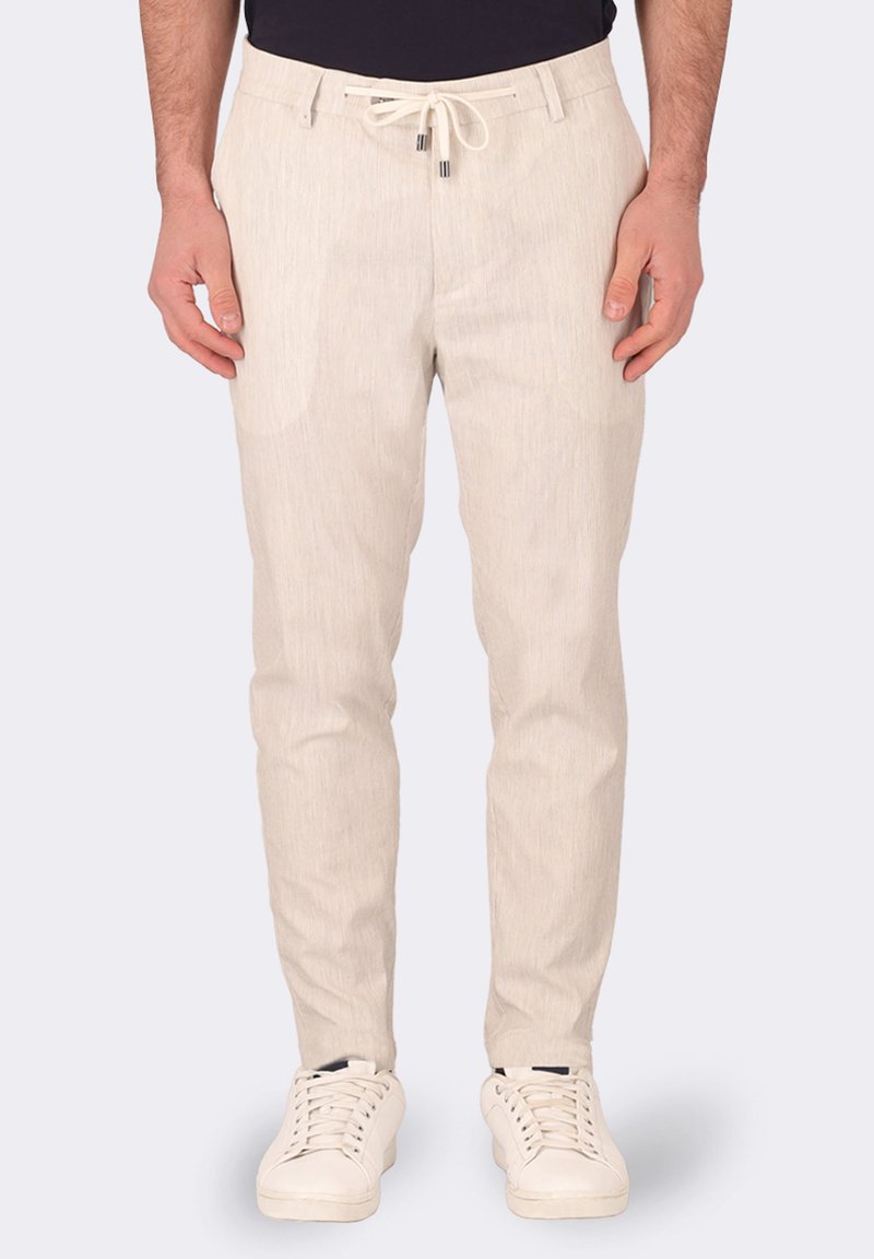Pantaloni beige chiaro realizzati in un tessuto testurizzato, con vita regolabile tramite cordoncino, tasche laterali e un design slim fit. Indossati con sneakers bianche.