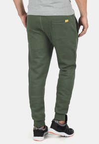 Green sweatpants with elastic cuffs, a single back pocket, and a yellow logo patch. Paired with black sneakers featuring white soles.
