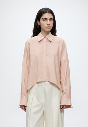 Joven mujer con cabello largo y oscuro, que lleva una camisa suelta de color rosa pálido con botones y pantalones blancos de cintura alta contra un fondo liso.