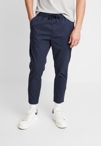 Homme portant un pantalon bleu marine raccourci avec cordon de serrage et des baskets blanches, debout devant un fond blanc uni.