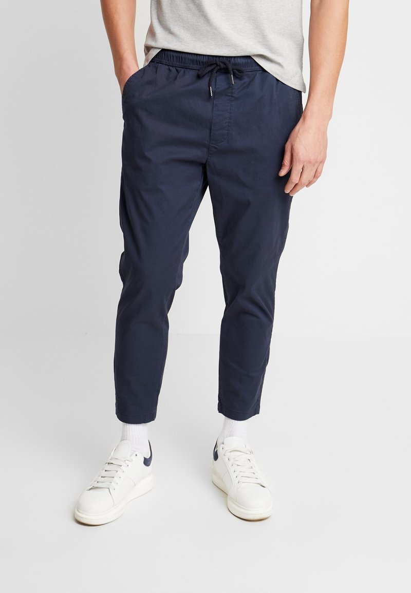 Homme portant un pantalon bleu marine raccourci avec cordon de serrage et des baskets blanches, debout devant un fond blanc uni.