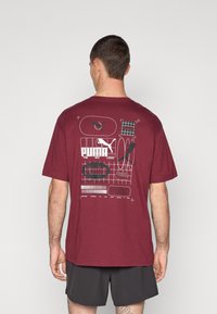 Maron farvet bomulds t-shirt med rund halsudskæring og korte ærmer. T-shirten har grafiske designs i hvid, grøn og rød på ryggen, inklusive logoer.