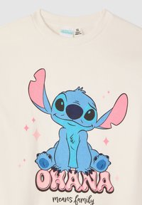 Maglietta bianca di cotone con un personaggio dei cartoni animati blu con grandi orecchie e occhi neri. Sotto il testo recita “OHANA” in rosa, con scintillii.