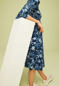 Vestido floral azul marino con un corpillo ajustado, falda acampanada, mangas tres cuartos y un objeto rectangular blanco sostenido a un lado.
