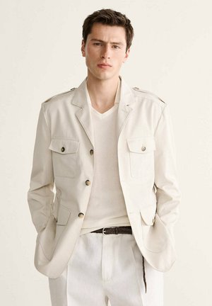 Joven con chaqueta beige de trabajo, camisa de punto blanca y pantalones claros con las manos en los bolsillos ante un fondo liso.