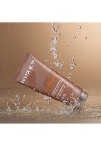 Brun tube av Nuxe Men Boost dushgel med vit och orange text, delvis nedsänkt i sprutande vatten mot en gradientbakgrund.