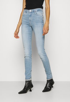 Kvinde iført lyseblå skinny jeans, sort ærmeløs top og sorte ankelstøvler med spids tå stående mod en hvid baggrund.