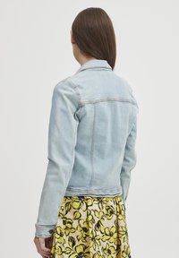 Šviesiai mėlyna džinsinė striukė su klasikiniais atlapais, ilgomis rankovėmis ir matomais siuvimo detalėmis, derinama su raštuota geltona gėlių sijonu.