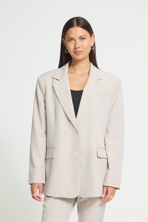 Femme aux longs cheveux foncés portant un blazer oversize beige clair et un pantalon assorti sur un haut noir, debout devant un fond uni.