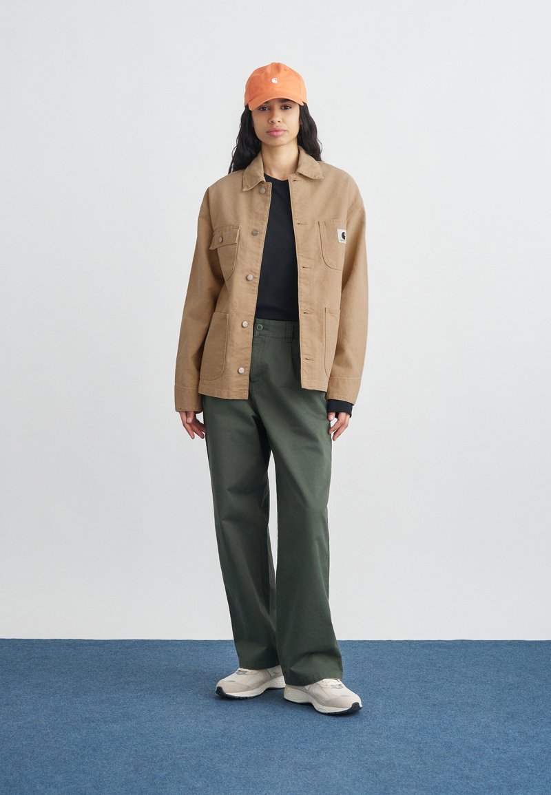 Jonge vrouw met een oranje pet, een bruine jas, een zwart shirt, olijfgroene broek en witte sneakers, staand op een blauw tapijt tegen een witte muur.