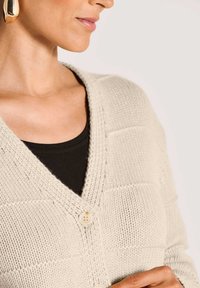 Frau in einer cremefarbenen gestrickten Strickjacke mit großen Knöpfen über einem schwarzen Top, mit sichtbarem goldenen Creolenohrring vor einem hellen Hintergrund.