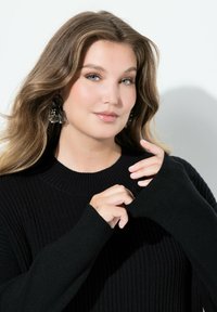 Svart ribbad stickad tröja med hög krage och förlängda ärmar. Modellen bär stora blommiga örhängen och håller i muddarna på tröjan.