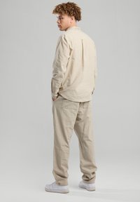 Camicia beige a maniche lunghe con bottoni e piega sul retro, abbinata a pantaloni larghi coordinati. Entrambi i capi sono realizzati in un tessuto morbido. Sneakers bianche.