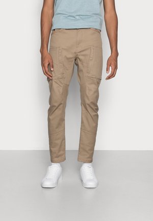 Mężczyzna w beżowych spodniach typu cargo i białych tenisówkach stoi na jasnym, jednolitym tle.