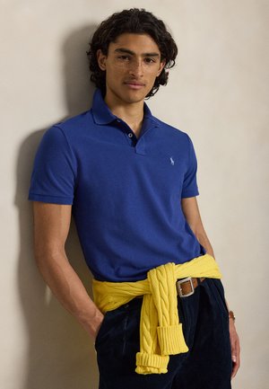 Ung person med krøllet hår iført en blå poloshirt og en gul sweater bundet om livet mod en ensfarvet væg.