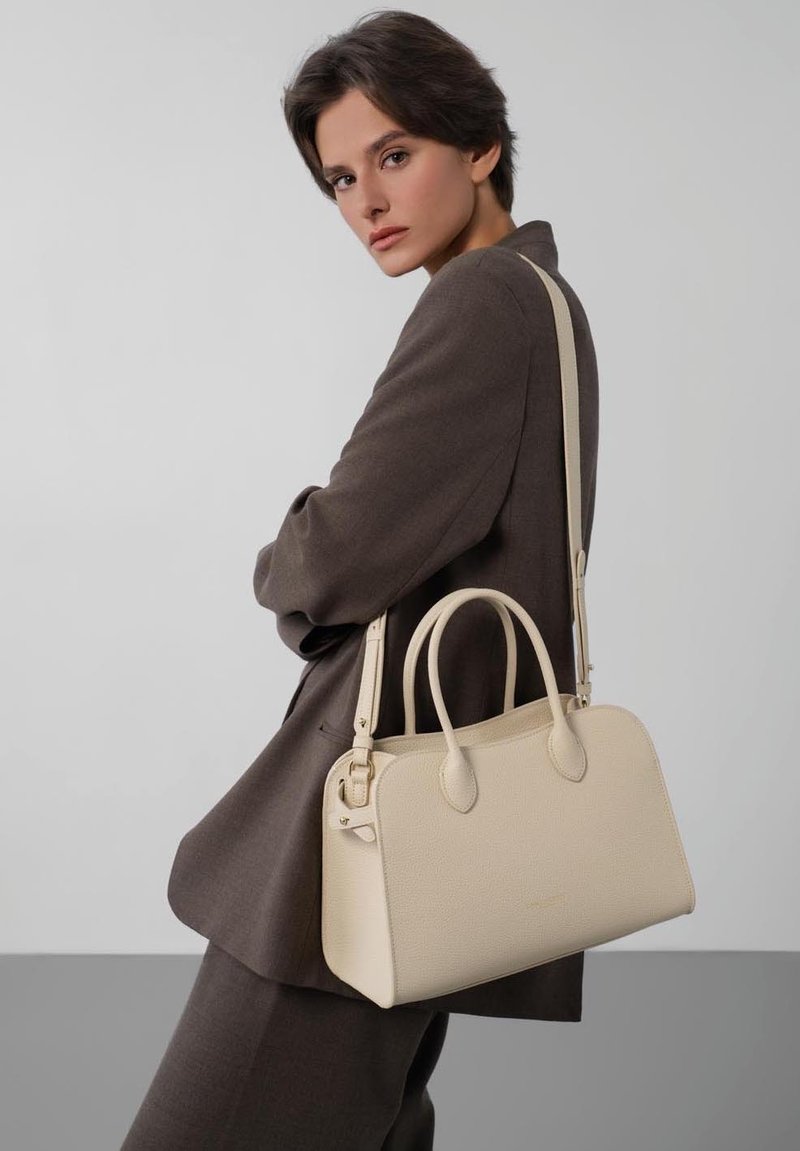 Femme portant un tailleur marron pose avec un sac à main en cuir crème suspendu à l'épaule sur un fond gris uni.