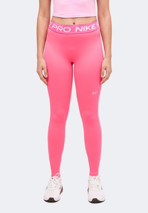 Kvinde iført lyserøde Nike Pro-leggings, lyserød crop top og hvide Nike-sportsko, stående med armene afslappet ned langs siden.