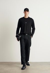 Zwarte shirt met lange mouwen met een kleine blauwe accent, zwarte broek en een zwarte pet. De figuur houdt een zwarte tas vast, staande tegen een neutrale achtergrond.