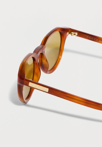 Στρογγυλά διαφανή γυαλιά ηλίου από ακετάτη κεχριμπαρένια-καφέ με φακούς με κίτρινη απόχρωση και χρυσαφένια πλακέτα με το λογότυπο Gucci στο πλαϊνό μέρος.