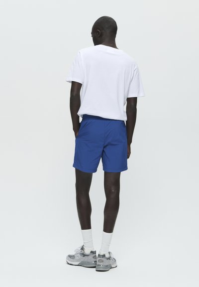 Blaue Shorts mit elastischem Bund, kombiniert mit einem weißen T-Shirt. Das Model trägt graue Sneakers und weiße Knöchelsocken. Minimalistisches Design.