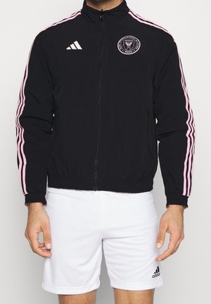 Homme portant une veste noire zippée avec des rayures roses et le logo de football de Miami, associée à un short blanc Adidas.