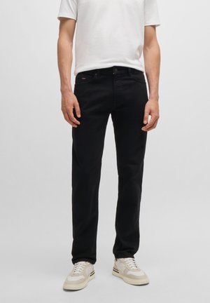 Mann trägt schmale schwarze Jeans, weißes kurzärmeliges T-Shirt und weiße Sneakers, steht vor einem einfarbigen hellgrauen Hintergrund.