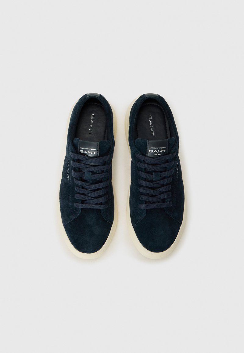 Un paio di sneakers in suede blu navale con suole bianche e lacci neri, marchiato GANT sulla linguetta e di lato, disposti in alto verso il basso su uno sfondo chiaro.