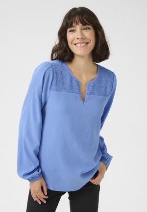 Blouse bleu clair avec un encolure smockée texturée, des manches bouffantes longues et un décolleté en V. Fabriquée en tissu doux avec une finition légèrement froissée.