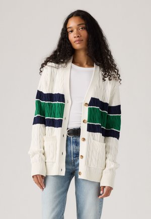 Vrouw in een witte kabelgebreide cardigan met marineblauwe en groene strepen over een witte top en lichtblauwe jeans, staand tegen een neutrale achtergrond.