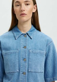Giacca di jeans azzurro chiaro con maniche corte, chiusura frontale con bottoni, due tasche sul petto e colletto classico. Presenta cuciture a contrasto.