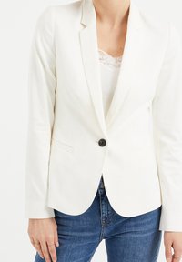 Vit skräddarsydd blazer med en enda svart knapp, slagkrage och två framfickor, som är kombinerad med en linne med spetsdetaljer och denimjeans.