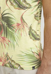 Camisa de manga corta en amarillo claro con un patrón de hojas de palma en verde y rosa. Tela suave y ligera. Incluye una pequeña etiqueta negra de la marca en el dobladillo.