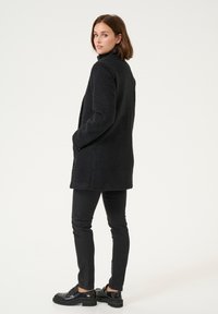 Zwart gestructureerd jasje met hoge kraag en zakken, gecombineerd met zwarte slanke broek en zwarte instappers, gezien van achteren.