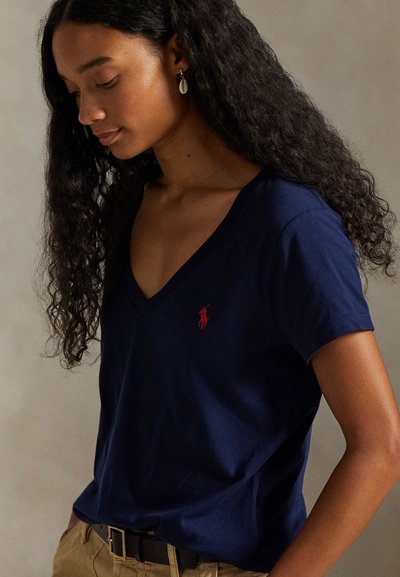 Γυναίκα με μακριά σγουρά μαλλιά που φοράει μπλε σκούρο T-shirt με V λαιμόκοψη και κόκκινο λογότυπο, μπεζ παντελόνια, μαύρη ζώνη και σκουλαρίκια σε σχήμα κοχυλιού, κοιτάζοντας προς τα κάτω.