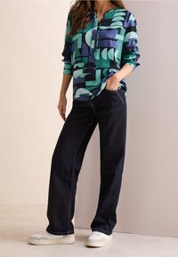 Chemise à manches longues avec un motif abstrait vert et bleu marine, associée à un pantalon large noir et des baskets blanches, offrant une tenue décontractée et confortable.