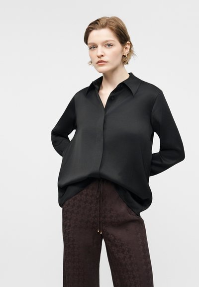 Γυναίκα με κοντά ανοιχτό καστανά μαλλιά, ντυμένη με φαρδιά μαύρη πουκαμίσα με κουμπιά και σκούρο καφέ παντελόνι με σχέδια, όρθια με τα χέρια πίσω από την πλάτη.