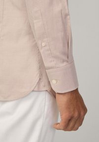 Chemise à carreaux marron et blanc avec des poignets à bouton, ourlet arrondi et texture subtile, portée sur un pantalon de couleur claire.