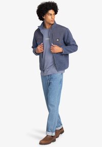 Navy fleece jacket with a smooth fabric front, two chest pockets, and a relaxed fit; paired with a gray tee and blue jeans.