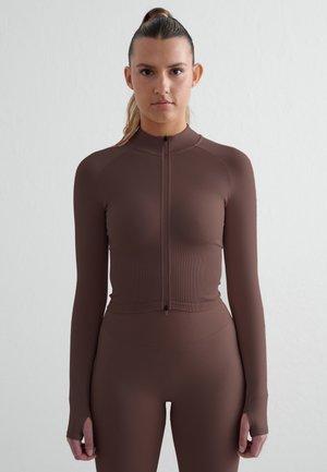 Donna con i capelli legati indossa una maglia marrone aderente a maniche lunghe con cerniera e leggings abbinati, in piedi davanti a uno sfondo chiaro uniforme.