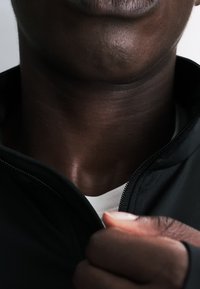Primer plano de la parte inferior del rostro y el cuello de una persona que lleva una chaqueta negra, sujetando la cremallera cerca de una camiseta blanca.