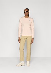 Langarm, lichtroze katoenen shirt met een klein blauw logo, gecombineerd met beige broeken en crèmekleurige sneakers. Accessoires zijn onder andere witte zonnebril.
