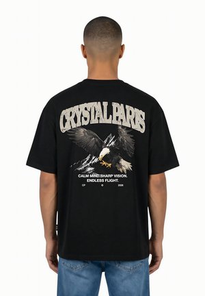 Uomo che indossa una maglietta nera con "CRYSTAL PARIS", aquila, montagne e testo "Mente calma. Visione nitida. Volo senza fine" sulla schiena.