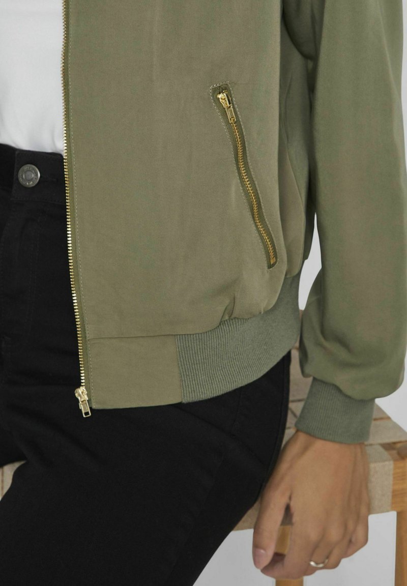 Olivgrüne Bomberjacke mit goldenen Reißverschluss und gerippten Bündchen, ausgestattet mit einer Seitentasche und glatter Textur, kombiniert mit schwarzen Hosen.