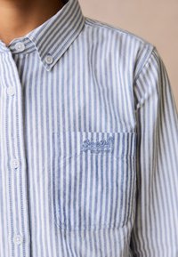 Világoskék és fehér függőleges csíkos ing, hegyes gallérral és egy kis zsebbel, amelyen hímzett márkajelzés található.