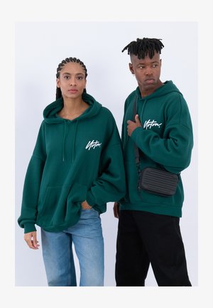 Zelené mikiny s kapucí a předními kapsami, volný střih. Jeden model nosí černé džíny, druhý nese černou crossbody tašku.