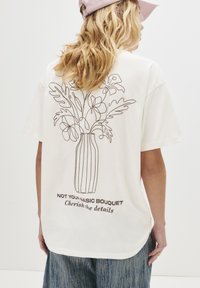 En blond person iført en hvid T-shirt med en strtegnet blomsterkrukke og teksten "Ikke din almindelige buket, værdsæt detaljerne" på ryggen.