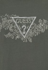 Haft logo GUESS w kształcie trójkąta z znakiem zapytania, z napisem "U.S.A." oraz "washed jeans" na ciemnym materiale z kwiatowym haftem.