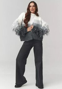 Sweter dzianinowy z teksturą w gradientowych odcieniach szarości, z frędzlami na rękawach; zestawiony z czarnymi spodniami z wysokim stanem i płaskimi czarnymi butami.